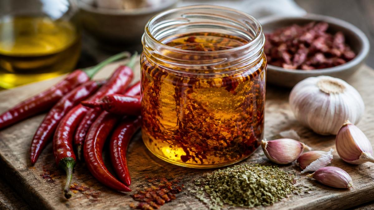 Glass jar of golden infused chilli oil with fresh garlic and dried herbs on a rustic wooden board