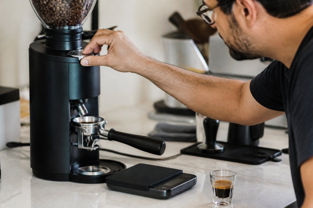 Close-up of a home barista's hands adjusting the grind dial on a burr grinder, with a portafilter and digital scale on the counter beside a freshly pulled espresso shot in a small glass