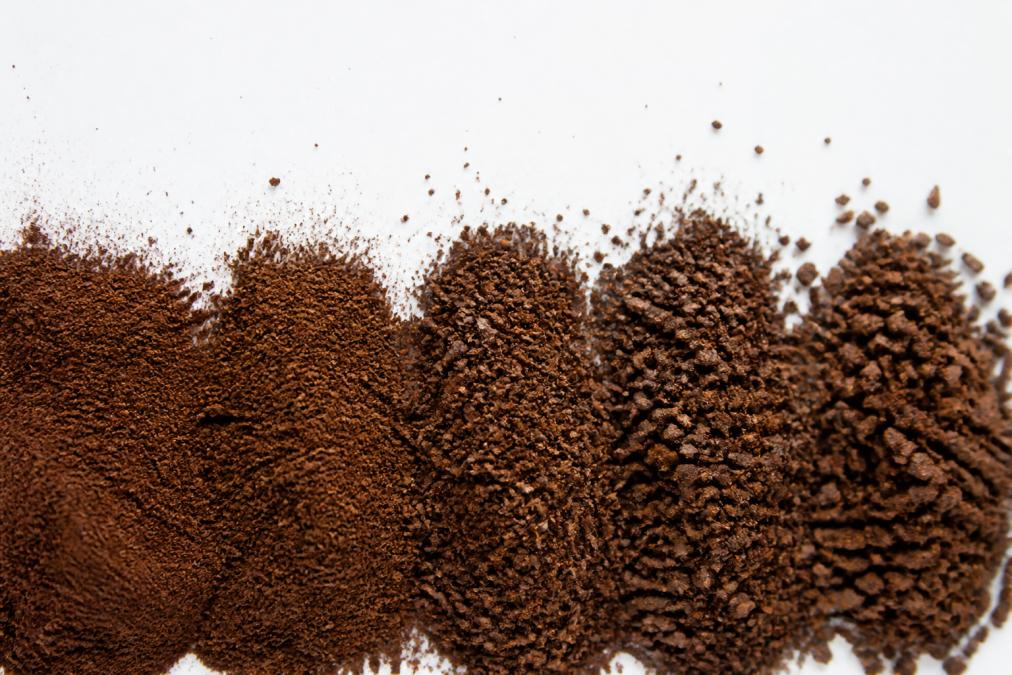 Close-up macro photography of espresso grind particles at different sizes, from fine powder to coarser granules, showing particle size distribution and consistency across a white surface under bright lighting