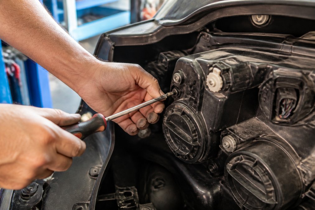 Mechanic's hands adjusting a headlight adjustment screw with a Phillips screwdriver on the rear of a vehicle headlight housing, close-up view showing the screw mechanism