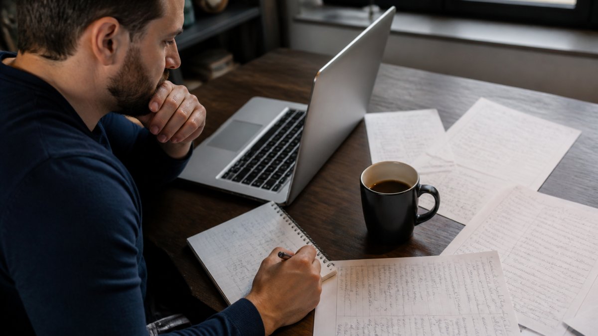 Professional analyst working at desk with laptop displaying spreadsheet data, coffee cup, and handwritten notes on multiple sheets of paper in focused workspace