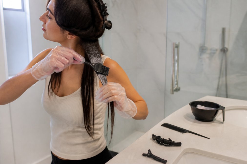 Woman wearing gloves applying at-home keratin treatment to hair sections with a brush in a bright bathroom