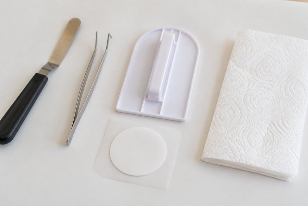 Flat lay of edible cupcake topper application tools arranged on white surface: small offset spatula, tweezers, silicone smoothing tool, paper towels, and a single edible topper with backing sheet visible