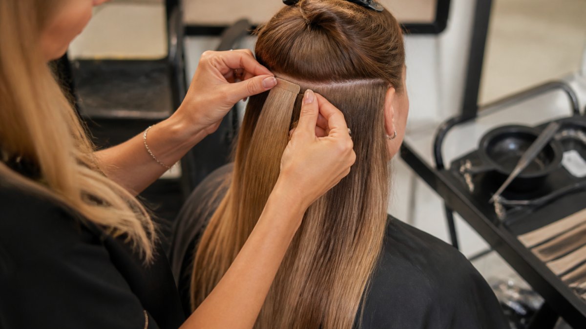 Close-up of a stylist's hands pressing a tape-in hair extension panel into a clean horizontal section of a client's hair, with a tail comb and sectioning clips visible on a salon trolley nearby