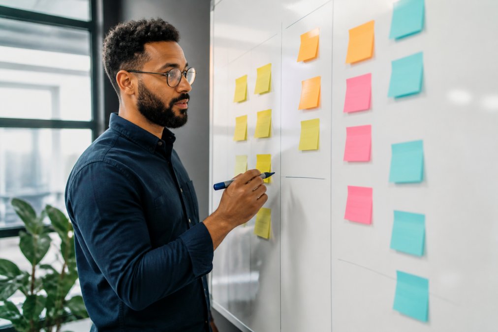 A founder standing at a whiteboard mapping out a 90-day timeline with sticky notes in three color-coded columns, holding a marker while reviewing milestones in a bright office