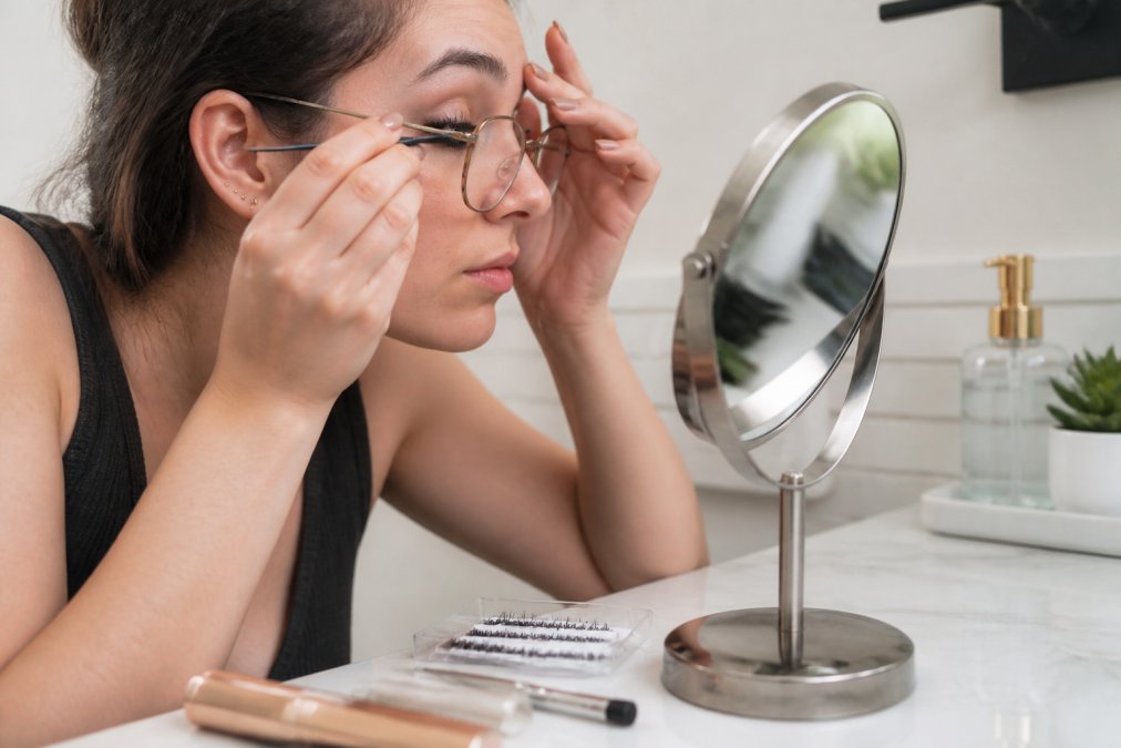Woman wearing glasses using curved tweezers to apply press-on lashes in front of a magnifying mirror