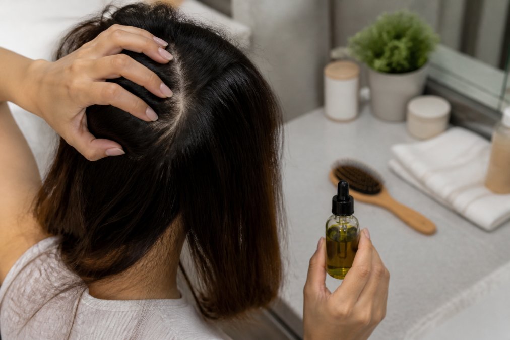Close-up of a woman's hands parting her hair at the crown with a dropper bottle of botanical serum nearby on a bathroom counter, soft natural light