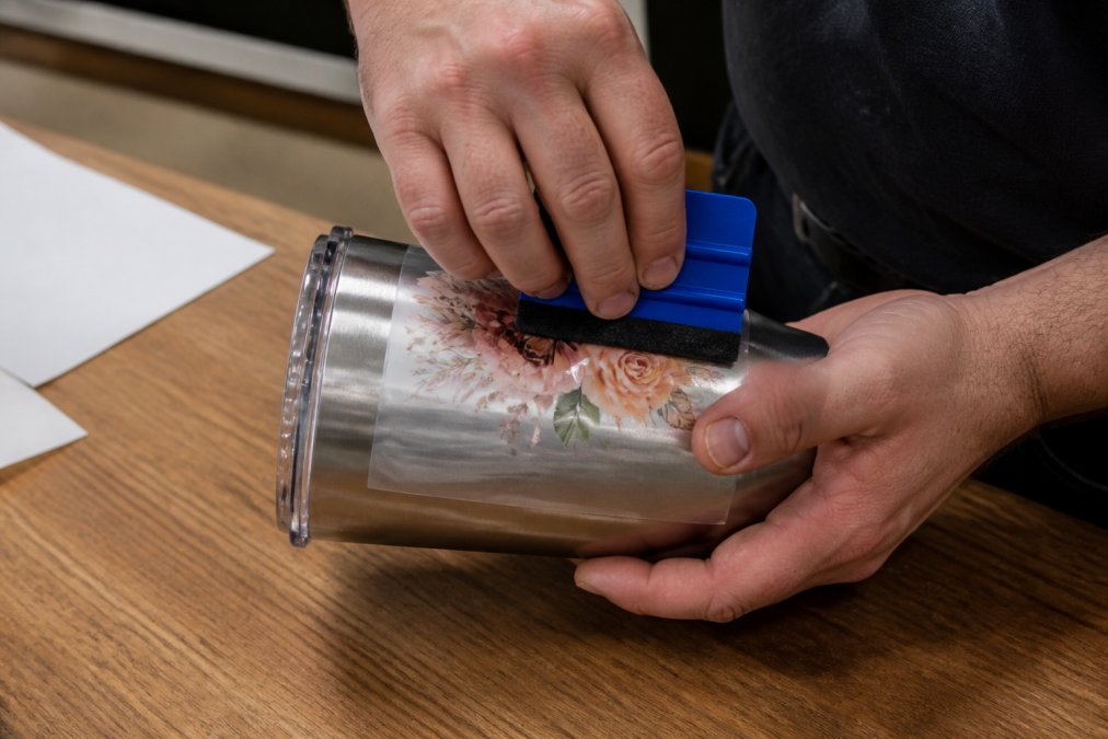 Close-up of hands in a bright workshop pressing a UV DTF decal onto a curved stainless steel tumbler with a squeegee, working from the centre outward