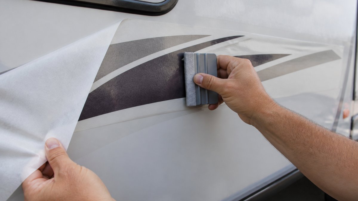 Close-up of hands using a grey felt-edge squeegee to smooth a large vinyl stripe decal onto the side panel of a caravan, backing paper partially peeled back and transfer tape visible, soft shaded daylight