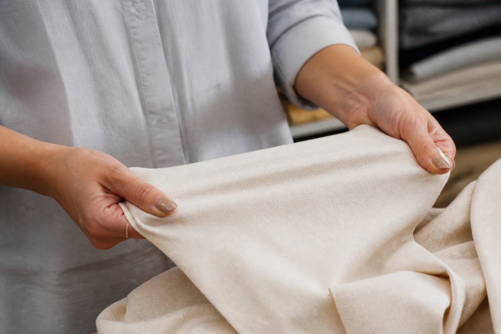 Close-up of a designer's hands performing a stretch test on a piece of natural cotton fabric, showing the weave structure and slight tension under bright workroom lighting