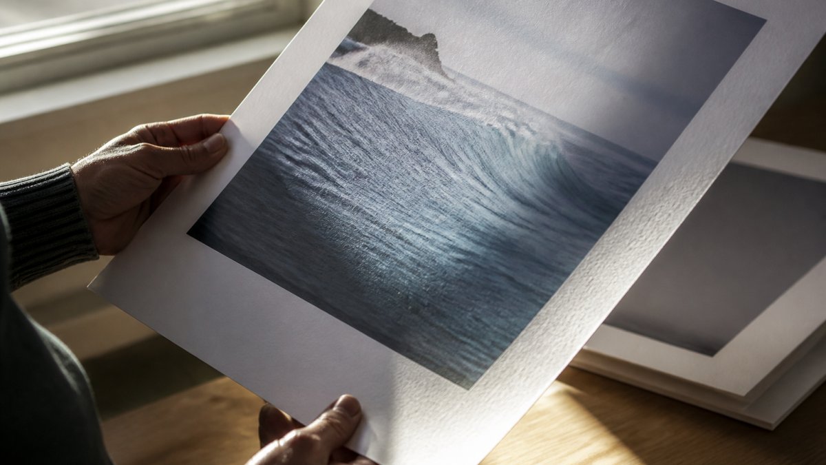 A close-up photograph of a collector's hands holding a fine art print at an angle near a window, catching raking light across the paper surface to reveal texture and depth of ink
