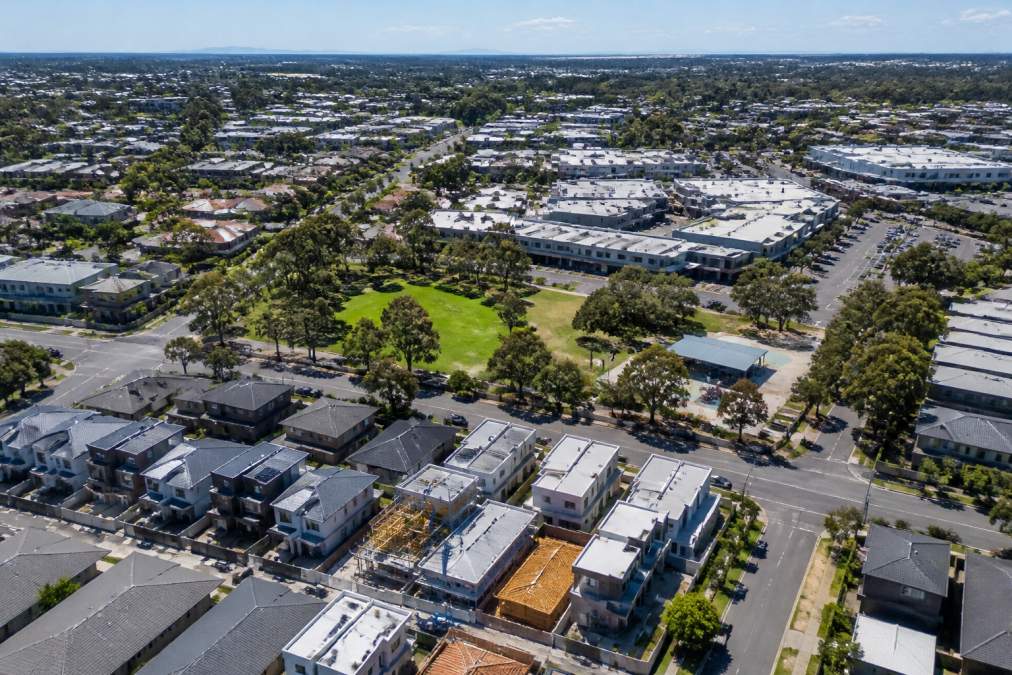 Aerial view of a growing suburban neighbourhood with new residential developments, modern shopping centres, parks and tree-lined streets under bright daylight