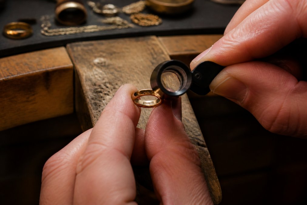 A jeweler uses a loupe to inspect a gold ring's hallmark stamp to verify jewelry quality on a workbench.