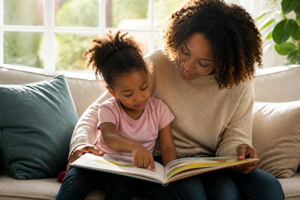 Parent and young child sitting together on a couch reading a colorful picture book, child's small hand resting on an open page with bright illustrations, natural afternoon sunlight streaming through a window behind them