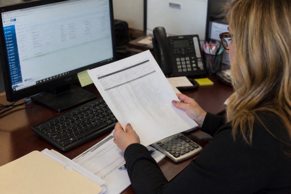 Office manager reconciling dealership vendor invoices by comparing printed statements to accounts payable software.