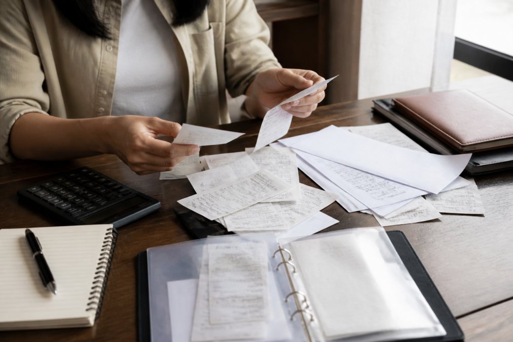 Close-up of hands organizing receipts and financial documents on a wooden desk with a calculator, notebook, and pen nearby under natural window lighting
