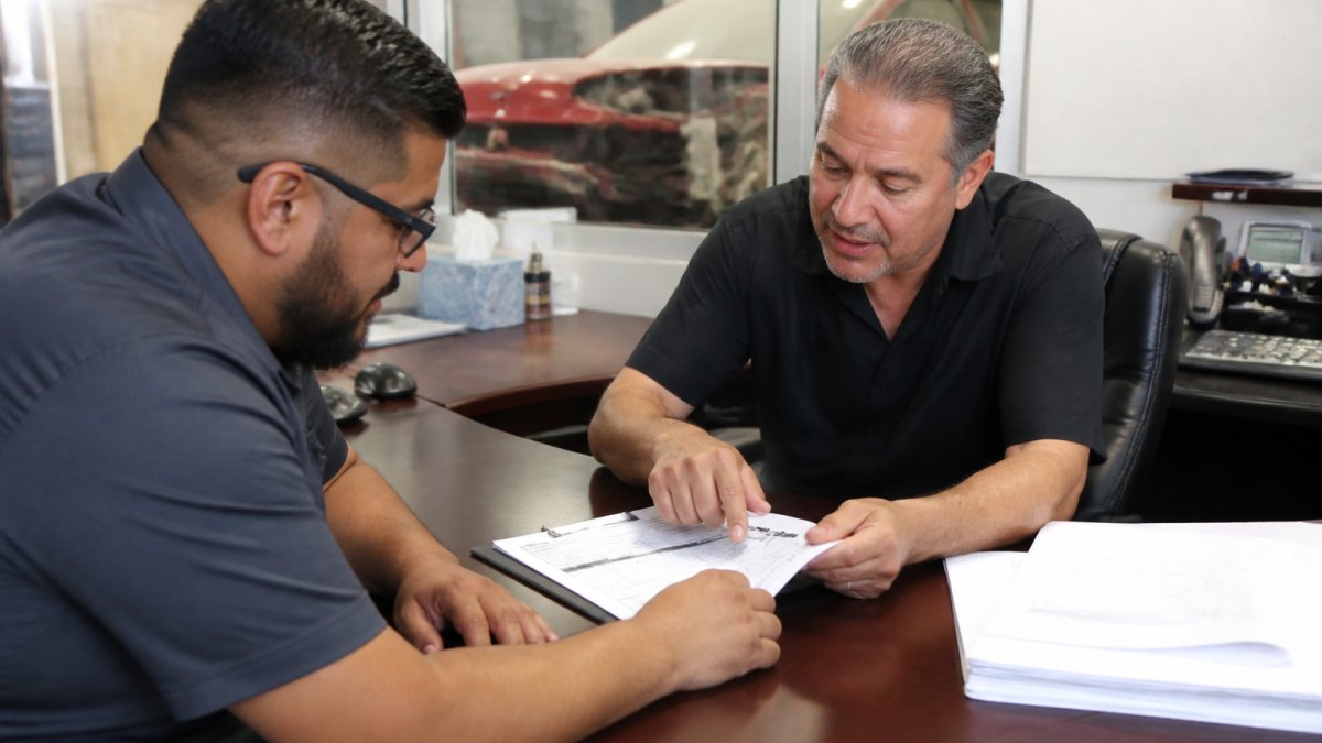 Professional service advisor sitting across from customer at desk in modern repair shop, reviewing written estimate document together while customer points at line items and asks questions, natural office lighting