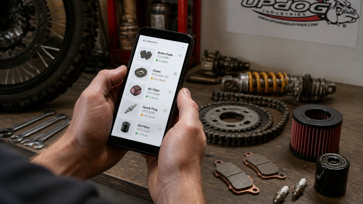 Close-up of hands holding a smartphone displaying a parts catalog interface with inventory status indicators, motorcycle workbench with tools and components visible in soft workshop lighting