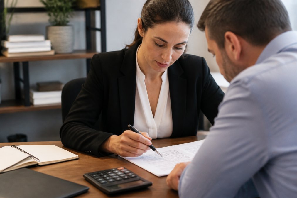 Professional in business attire sitting at desk with loan documents, calculator, and notepad, reviewing paperwork with focused attention under office lighting