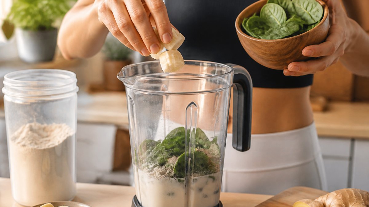 Close-up of hands adding fresh banana, spinach, and ginger to a blender with plant-based protein powder and warm water, natural kitchen lighting