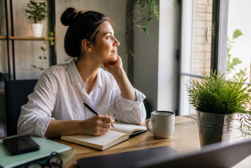 Person sitting peacefully at a desk with a journal, coffee, and plant, taking a mindful break during a work day with soft natural lighting filtering through a window