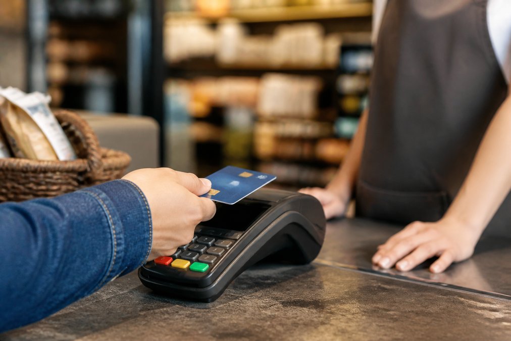 Close-up of a person tapping a contactless card on a modern payment terminal at a retail counter, with blurred shelves and warm store lighting in the background