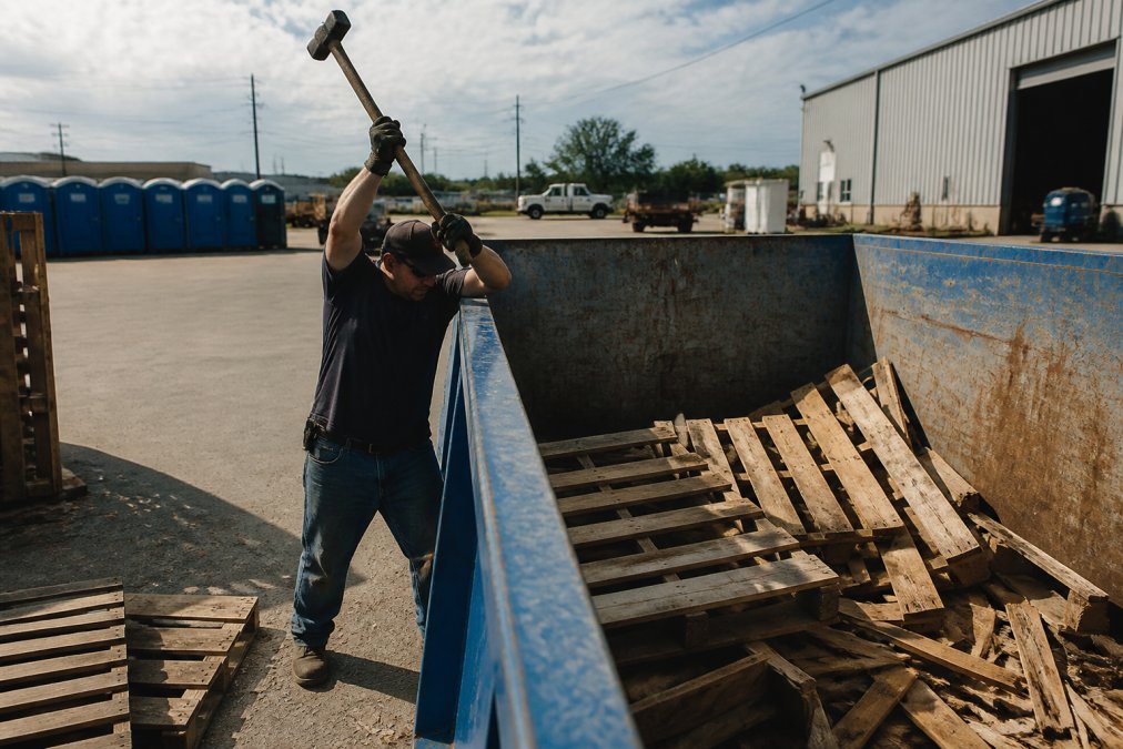 Worker breaking down wooden pallets with a sledgehammer over a roll-off dumpster, demonstrating proper technique for reducing bulky item size with controlled overhead swing