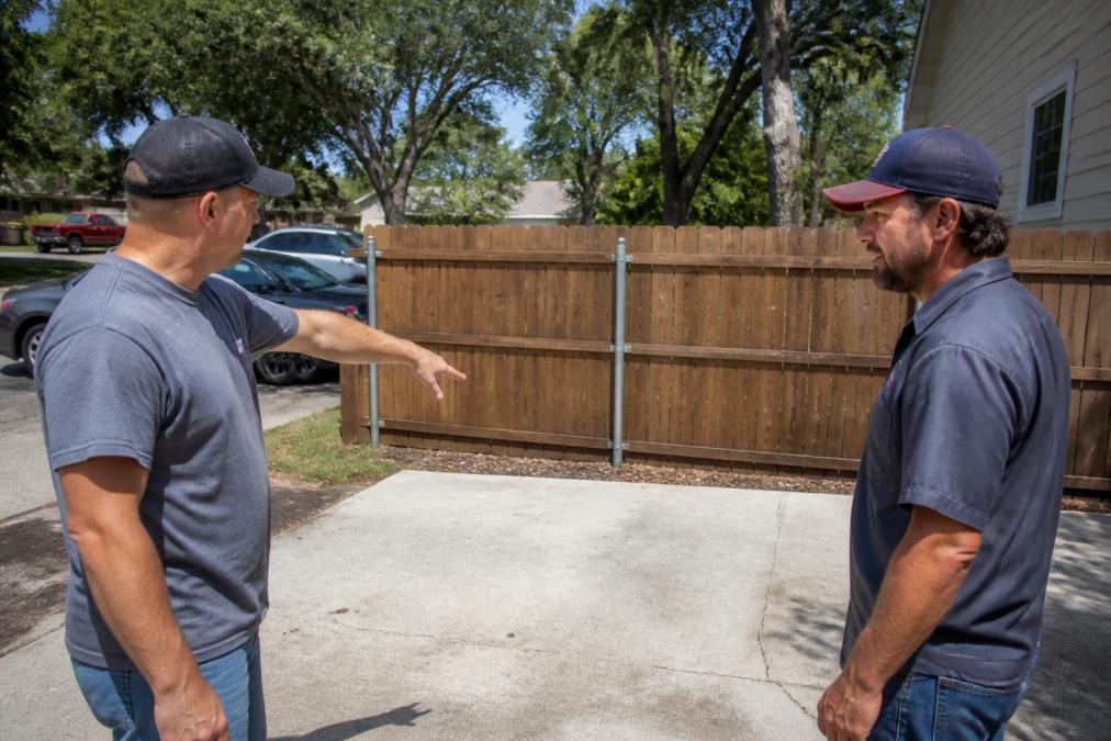 A homeowner and a dumpster delivery driver standing in a residential driveway, pointing at a cleared concrete pad where a roll-off dumpster will be placed, with a wooden fence and parked cars visible in the background