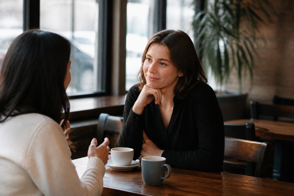 Two people having a genuine conversation over coffee in a warm, naturally lit café setting, one person leaning forward with engaged expression while the other speaks, suggesting active listening and genuine connection