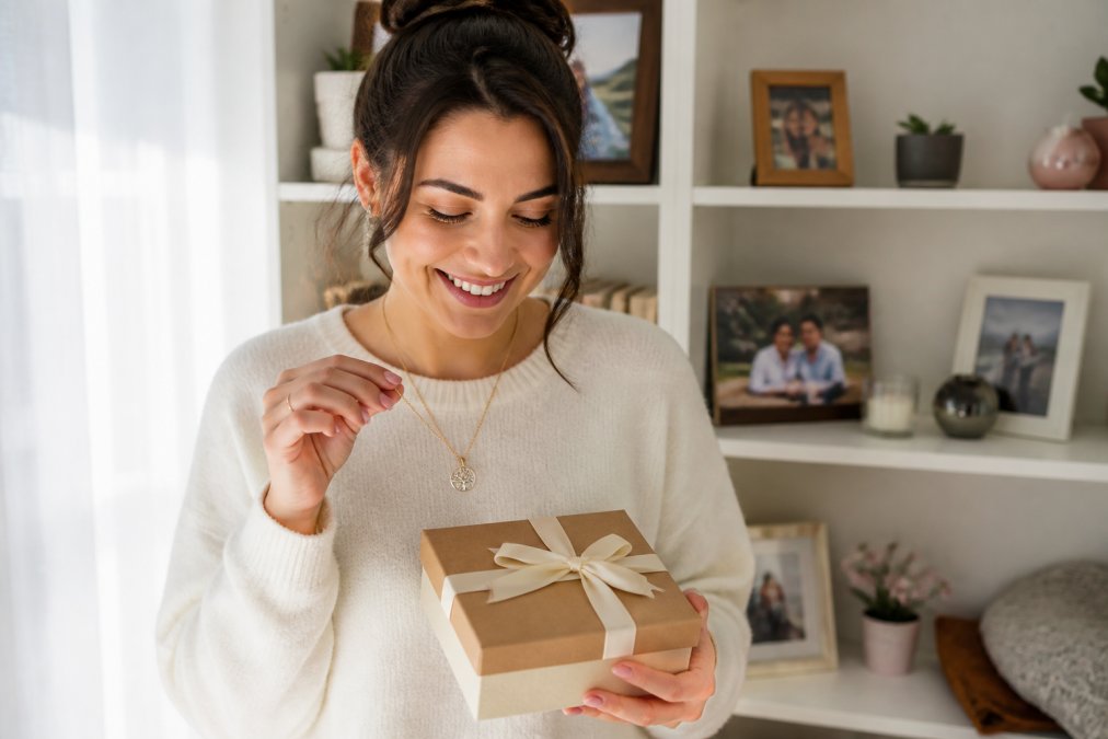 A woman holding a personalized necklace and thoughtfully wrapped gift box, smiling warmly while surrounded by meaningful keepsakes and photos on a shelf