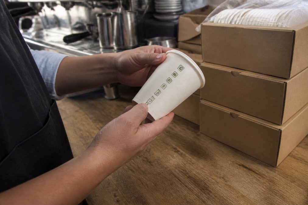 Close-up of a cafe owner's hands inspecting the certification logo printed on the side of a compostable paper takeaway cup, with a stack of unopened packaging boxes on the counter behind