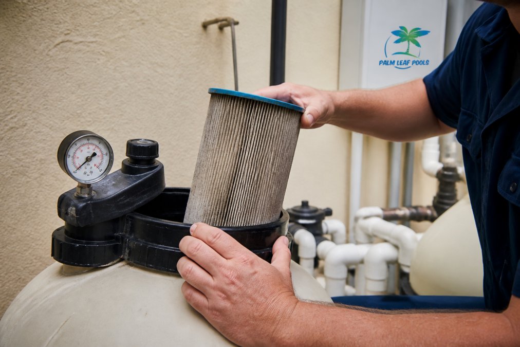 Close-up of a pool filter system showing a pressure gauge reading high, with a technician's hand checking the cartridge condition and examining filter housing