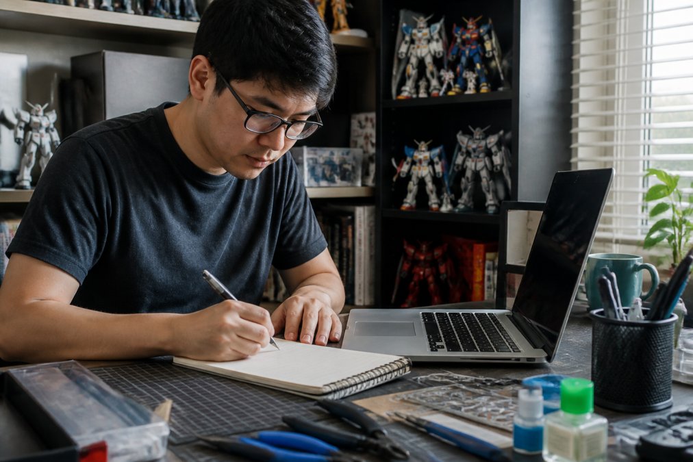 A collector at a desk writing a monthly hobby budget in a notebook, with a laptop showing a spreadsheet and a few collectible figures on a shelf nearby
