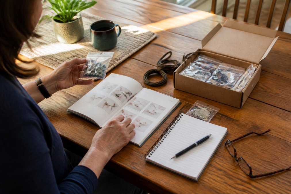 Person sitting at a wooden table examining craft kit contents spread out with instruction manual, material checklist written on notepad, and reading glasses nearby in natural afternoon lighting
