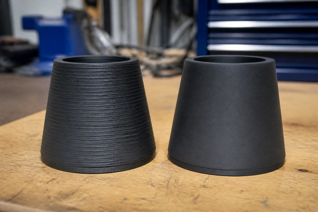 Close-up of two 3D printed parts side by side on a workbench, one with visible ridged layer lines and one with a noticeably smoother surface finish, both under bright workshop lighting