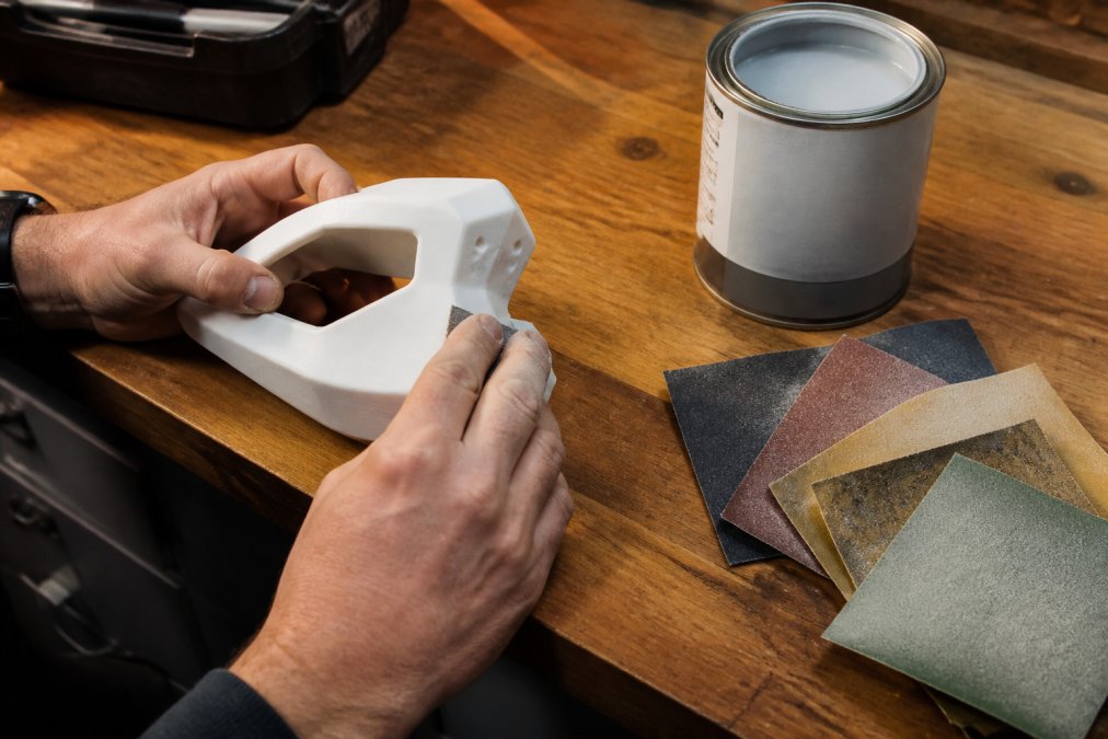 Hands sanding a white 3D printed part on a wooden workshop table under warm overhead lighting, with several sheets of sandpaper at different grits and a can of filler primer visible nearby