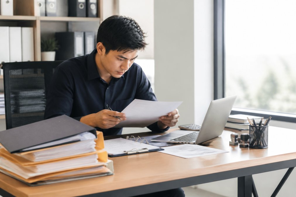 Procurement professional reviewing supplier documentation, certificates, and business records at desk with multiple folders, laptop, and compliance checklists in modern office with natural window lighting