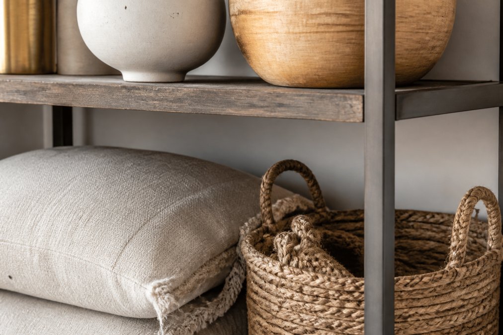 Close-up detail of a styled shelf corner showing mixed materials and finishes, soft linen fabric, warm wood texture, brushed metal frame, smooth ceramic vessel, and woven natural fiber basket creating visual depth through varied surface finishes and tactile contrast