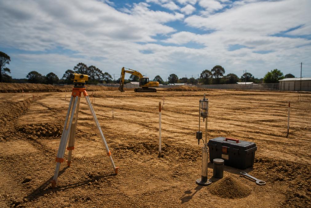 Wide shot of a construction site with surveying equipment, soil testing apparatus, and site markers visible on cleared land ready for foundation work, showing the scale of site preparation