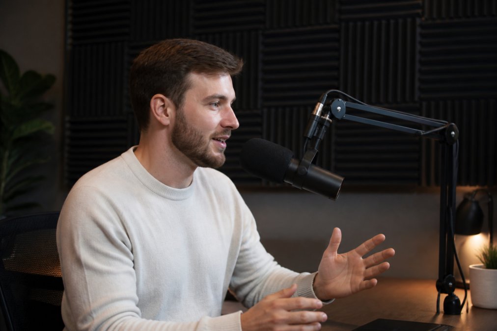 Content creator positioned at desk with microphone mounted on arm, acoustic foam panels visible on wall behind, proper microphone distance from mouth demonstrated with clear sight lines