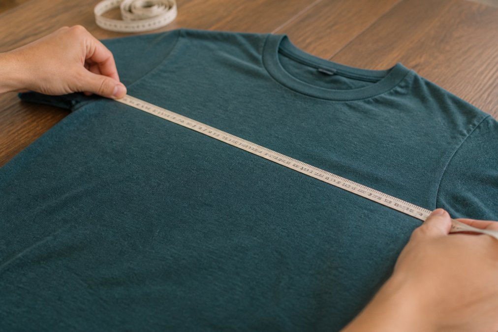 Close-up of a soft measuring tape stretched across a fitted T-shirt laid flat on a table, showing chest measurement clearly marked with fingers at the tape