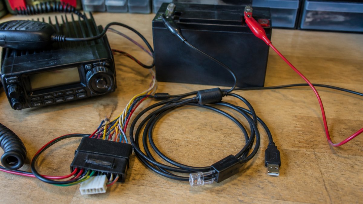 Close-up of radio equipment with wiring harness, 12V battery terminals, and programming cable clearly visible on a workbench with proper labeling and safety markers