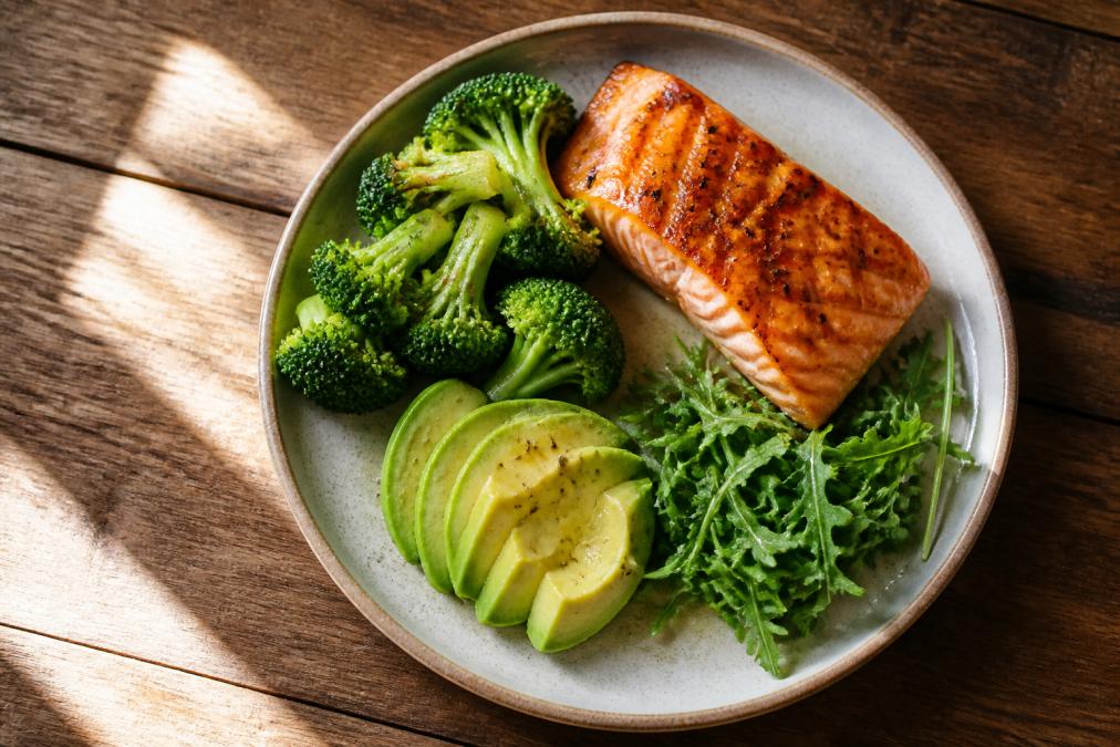 Overhead shot of a colorful plate with grilled salmon, steamed broccoli florets, sliced avocado, and fresh arugula on a wooden table with natural morning sunlight streaming across the surface