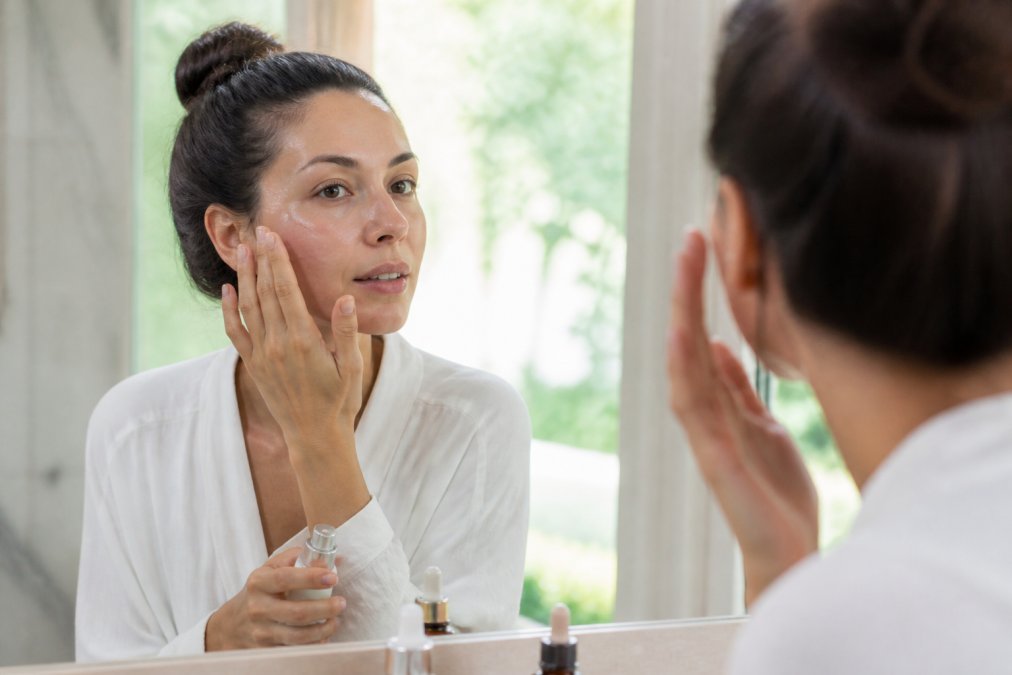 A woman in her 30s applying a light serum to her cheek with a fingertip, looking into a mirror in a bright, spa-like bathroom setting with soft natural lighting