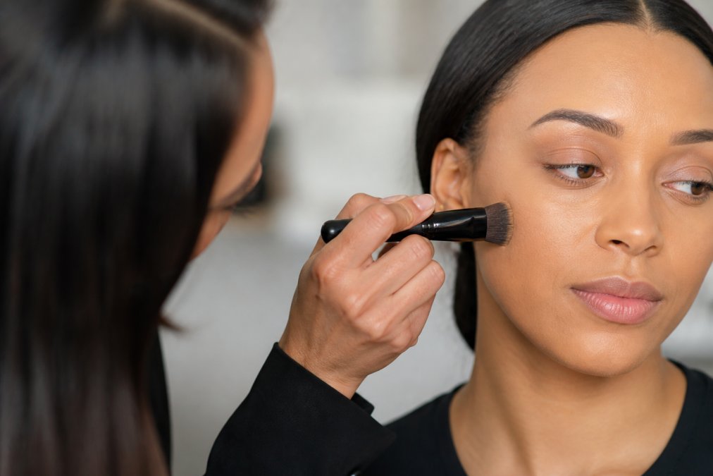 Close-up of a make-up artist's hand holding a professional foundation brush, applying foundation to a model's cheek with a smooth, circular motion against a clean studio background, with soft diffused lighting