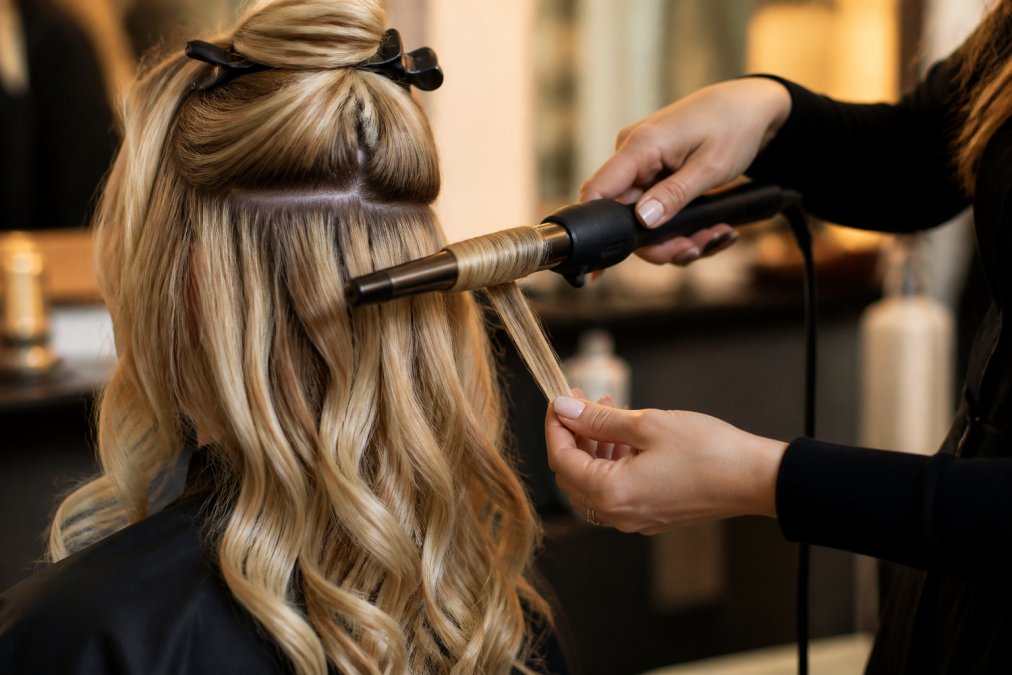 Hands using a curling wand on both natural hair and extensions simultaneously, creating unified waves and texture, with warm studio lighting highlighting the blended result