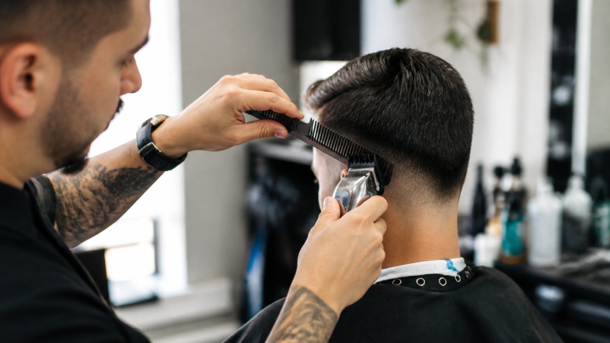 Professional barber demonstrating clipper-over-comb blending technique with client, showing upward hand motion and proper comb angle under natural barbershop lighting