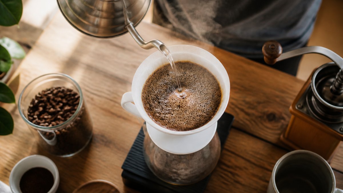 Overhead close-up of a gooseneck kettle pouring a thin stream of hot water over dark coffee grounds in a white ceramic pour-over dripper, bubbles and steam rising from the wet coffee bed, warm morning light on a wooden counter