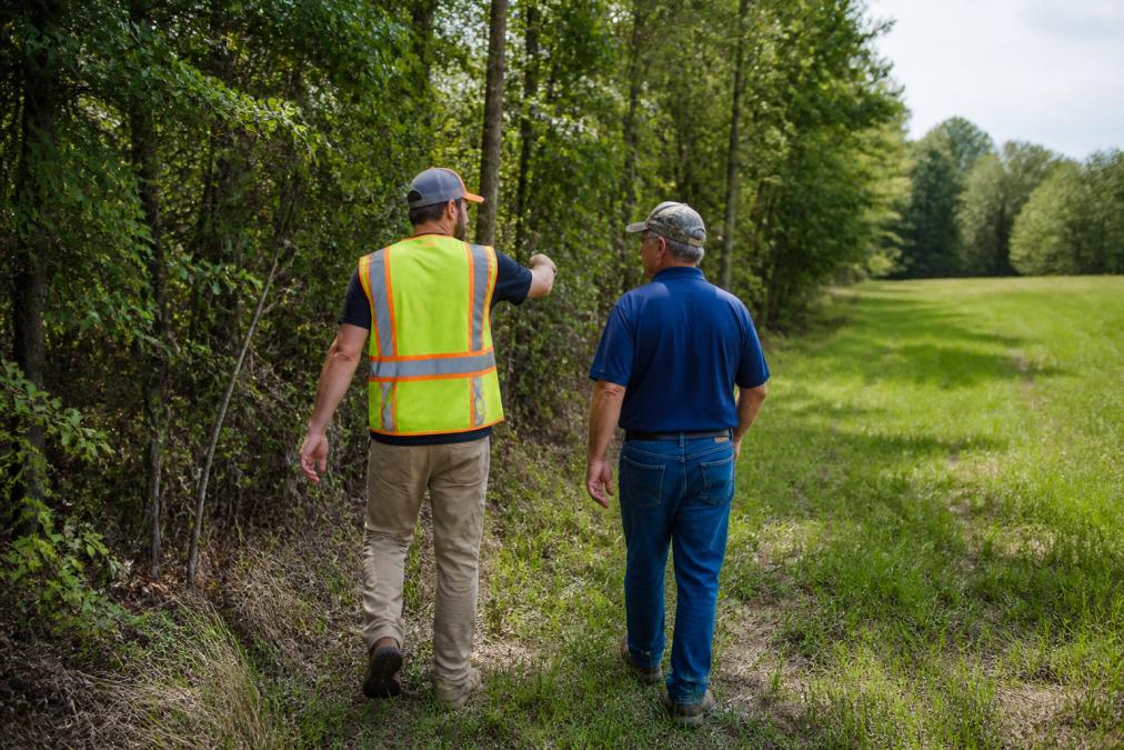 A land clearing professional in a high-visibility safety vest walking a wooded property boundary with a landowner, both examining the overgrown tree line during a site assessment visit in afternoon light