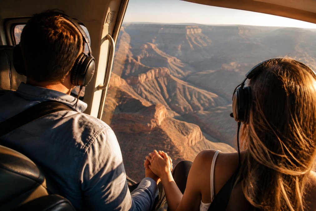 Couple holding hands in open door of helicopter with expansive aerial canyon views and golden-hour sunlight illuminating the landscape below